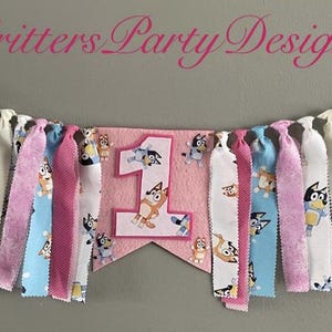 May include: A colorful fabric banner with the text "Critters Party Designs" and a pink pennant with the number "1" and cartoon characters. The banner features alternating strips of pink, blue, and white fabric.