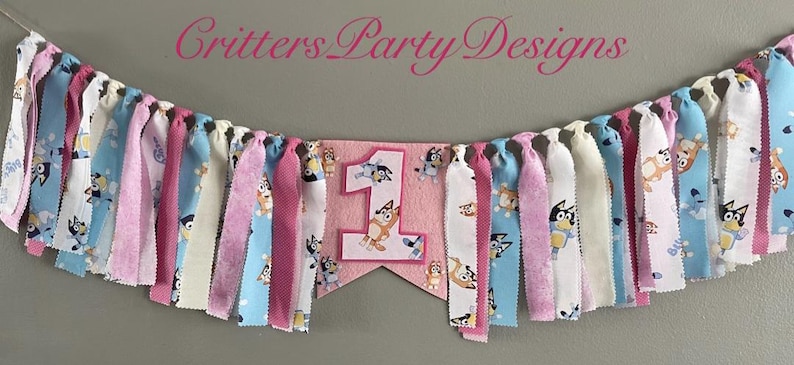 May include: A colorful fabric banner with the text "Critters Party Designs" and a pink pennant with the number "1" and cartoon characters. The banner features alternating strips of pink, blue, and white fabric.