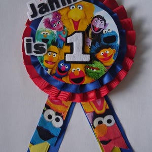 May include: A circular fabric rosette badge featuring a central image of Sesame Street characters. The badge has red and blue pleated ribbon trim, white felt lettering reading Jahmir is 1, and two hanging ribbon tails with character prints.