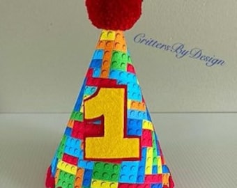 May include: A red and yellow birthday party hat with a red pom-pom on top. The hat is decorated with a pattern of colorful building blocks and the number "1" in yellow felt. The hat has a red scalloped edge and a white string.