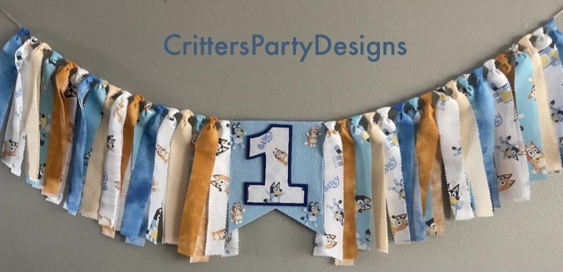 May include: A fabric banner with blue, white, and tan ribbons. The banner has a light blue pennant with a white number 1 and a cartoon dog. The text "CrittersPartyDesigns" is above the banner.