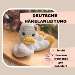 Könnte beinhalten: Ein gehäkeltes Amigurumi-Spielzeug in Form einer Ente mit gelbem Schnabel und einer rosa Blume auf dem Kopf. Das Spielzeug liegt auf einer Holzoberfläche. Das Bild enthält den Text "DEUTSCHE HÄKELANLEITUNG" und eine Liste von Merkmalen: "leicht", "druckerfreundlich", "gut bebildert".