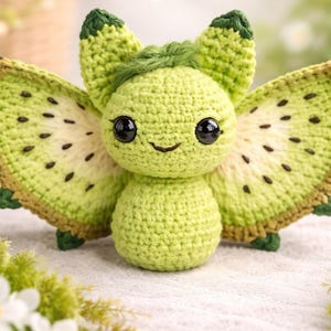 May include: A handmade crochet creature in shades of green, resembling a whimsical insect or fairy. It features large, detailed wings with a kiwi fruit design, black eyes, and a smiling face. The craftwork is set against a soft, blurred background.