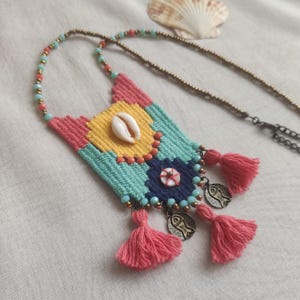 May include: A colorful woven necklace with a seashell pendant, pink tassels, and metal charms. The necklace is made with a variety of colors, including pink, yellow, blue, and green.