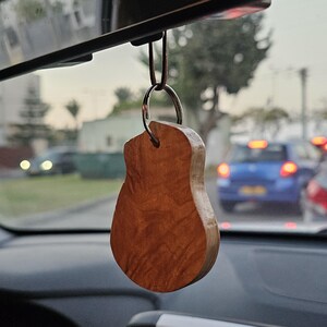 May include: A wooden guitar-shaped car charm hanging from a rearview mirror. The charm is a light brown color with a wood grain pattern.