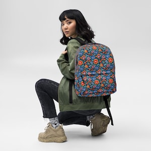 May include: A backpack featuring a colourful floral design in blue, orange, and red hues. The backpack has black straps and a black zip. The bag is on the back of a person wearing a green jacket and black jeans.