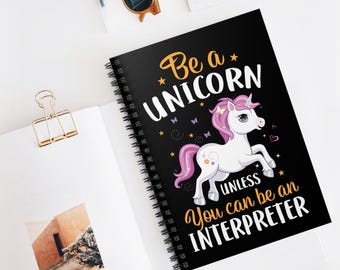 以下が含まれることがあります： 黒い表紙にユニコーンのイラストと「Be a UNICORN unless You can be an INTERPRETER」の文字が入ったスパイラルノート。ユニコーンはピンクのたてがみと尾を持っています。ノートは白い表面に開かれています。