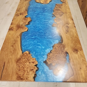 May include: A rectangular wooden coffee table with a blue epoxy resin river design. The wood has a natural grain and burl pattern, with the blue resin flowing through the center. The table is approximately 72 inches long.
