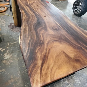 May include: A large, dark wood table top with a live edge and a natural wood grain pattern. The table top is supported by a black metal base.