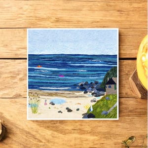 May include: Square art print showing a coastal scene. The artwork features a blue ocean with two small boats, a sandy beach, and a small house on a green hillside. The sky is light blue.
