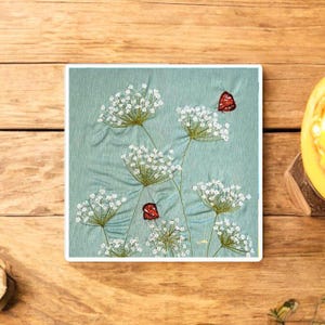 May include: Embroidered artwork in a square frame, depicting white flower heads and red butterflies on a pale blue fabric. The piece is set on a wooden surface. The image is a close-up of the embroidery.
