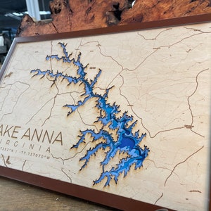 May include: A wooden map of Lake Anna, Virginia, with a brown frame. The lake is depicted in shades of blue, and the surrounding area is shown in beige. The text "LAKE ANNA VIRGINIA 38.027392°N -77.737070°W" is printed below the lake.