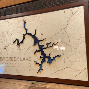 May include: A wooden map of Deep Creek Lake in Maryland, framed in brown wood. The lake is carved out of the wood and painted blue. The map is surrounded by a brown frame.