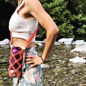 Könnte beinhalten: Eine Frau trägt ein weißes Tanktop und einen Blumenrock und trägt eine lila Wasserflasche in einem leuchtend orangefarbenen gehäkelten Trageriemen. Der Trageriemen wird über den Körper getragen und hat ein geknotetes Design.