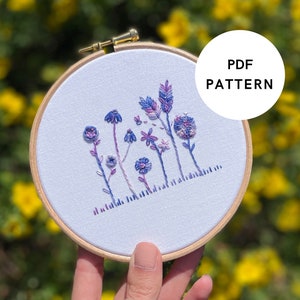 Può includere: Un primo piano di un cerchio da ricamo bianco con un disegno floreale ricamato in tonalità di viola e blu. Il cerchio è tenuto in una mano su uno sfondo sfocato di fiori gialli. Il testo "PDF PATTERN" è visibile in un cerchio bianco.