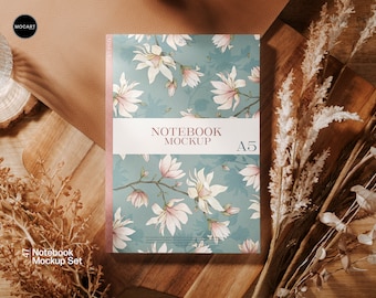 May include: A5 notebook mockup with a floral design featuring white and pink magnolia blossoms on a teal background. The notebook is labeled "NOTEBOOK MOCKUP" and includes the size "A5". The image is styled with dried plants and a glass of water.