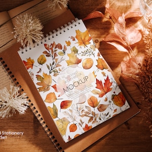 May include: A brown spiral bound notepad with a white cover featuring a watercolor design of autumn leaves in shades of orange, yellow, and red. The text "Notepad Sketchbook Mockup" is printed on the cover.
