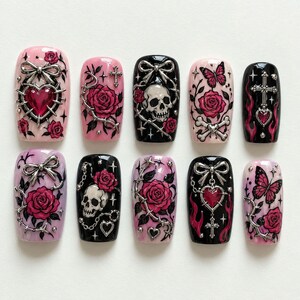 May include: A set of ten press-on nails featuring a gothic aesthetic with pink, purple, and black backgrounds. Designs include silver 3D bows, metallic hearts, skulls, roses, butterflies, crosses, and flame motifs, accented with small silver studs.