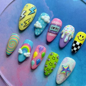 May include: A collection of ten colorful, decorated acrylic nails. Designs include lightning bolts, clouds, rainbows, balloons, a cassette tape with the word "GROOVY", sprinkles, a smiley face, a checkerboard pattern, and a star.