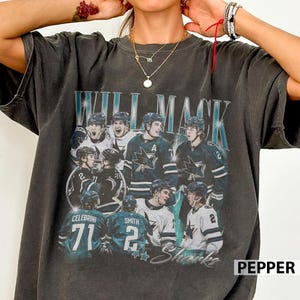 May include: A charcoal gray short-sleeve t-shirt featuring a vintage-style graphic of San Jose Sharks hockey players. The design includes the text WILL MACK, player names like CELEBRINI and SMITH, and various team jersey numbers in a collage layout.