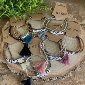 Puede incluir: Conjunto de siete pulseras de cuentas con diferentes palabras escritas en cuentas blancas, incluyendo "Fe", "Amor", "Gracia", "Valiente", "Humilde", "Amable" y "Agradecido". Cada pulsera tiene un pequeño borla.