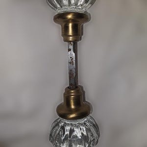 May include: A pair of clear, fluted glass door knobs connected by a central metal spindle. Each knob features a brass-toned metal base with a weathered finish. The central square metal rod shows signs of oxidation and rust along its length.