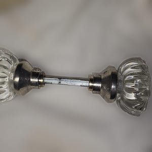 May include: A pair of clear, fluted glass door knobs connected by a central metal spindle. Each knob features a faceted, multi-sided design with a polished silver-tone metal base that attaches to the threaded square metal rod.