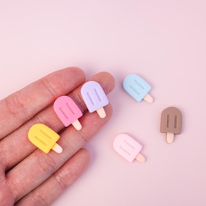 Puede incluir: Una colección de mini abalorios de helado de colores amarillo, rosa, morado, azul claro y marrón. Cada abalorio tiene un palito blanco. Los abalorios están dispuestos sobre un fondo rosa claro.