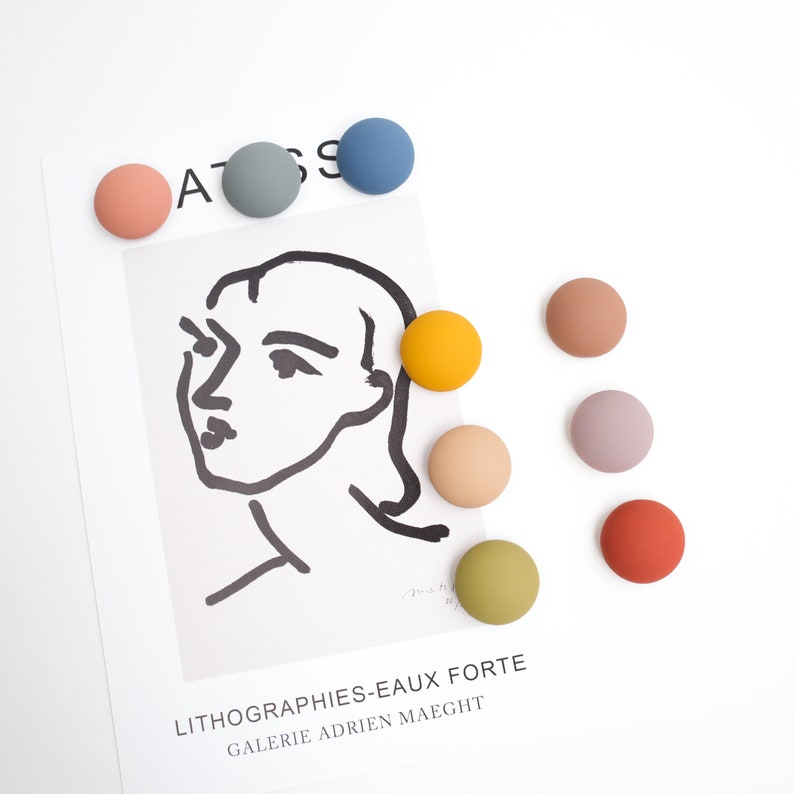Pu&ograve; includere: Assortimento di bottoni rotondi colorati in varie tonalit&agrave; di blu, giallo, arancione, marrone, verde e rosa. I bottoni sono disposti su una superficie bianca con un disegno in bianco e nero di un viso su uno sfondo bianco. Il testo "Lithographies-Eaux Forte Galerie Adrien Maeght" &egrave; stampato sotto il disegno.