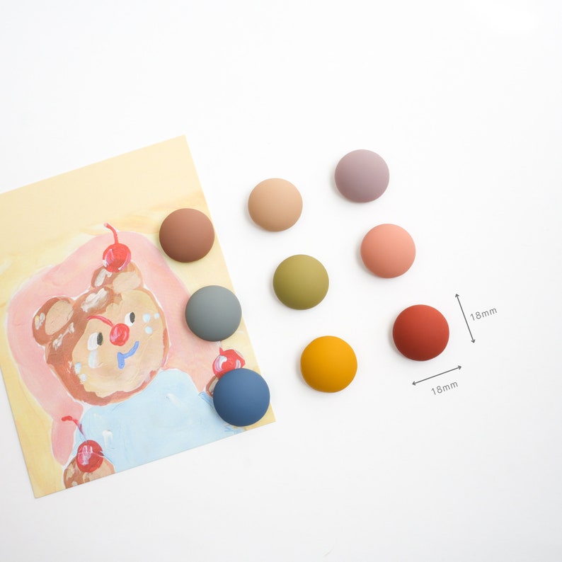 Pu&ograve; includere: Otto bottoni rotondi, piatti e colorati in ceramica da 1,8 pollici in varie tonalit&agrave; di marrone, verde, blu, arancione e rosa. I bottoni sono disposti in cerchio su una superficie bianca.
