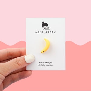 May include: A yellow banana shaped pin with black dots on a white card with the text "MINI STORY" and social media information.