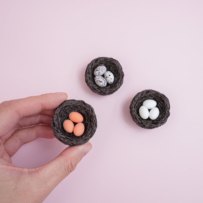 May include: Three small nests made of woven brown material, each containing different colored eggs. One nest has three orange eggs, another has three white eggs, and the last has three speckled eggs.