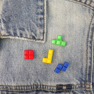 May include: A close-up of a denim jacket with four colorful plastic Tetris blocks attached to it. The blocks are red, yellow, green, and blue.