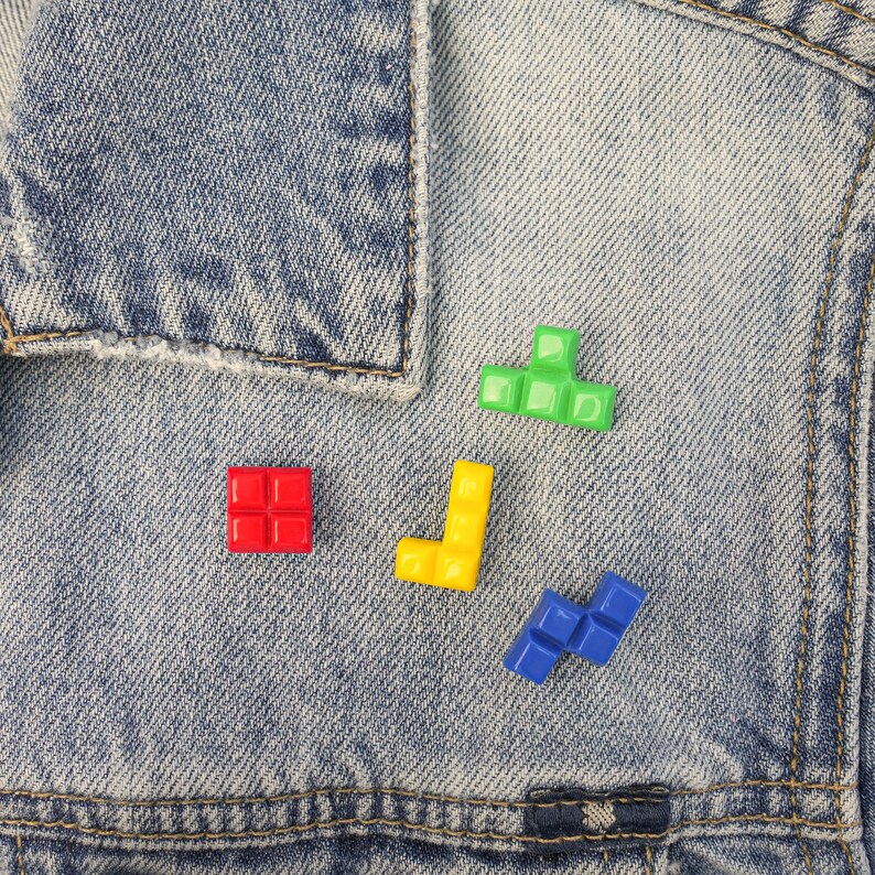 May include: A close-up of a denim jacket with four colorful plastic Tetris blocks attached to it. The blocks are red, yellow, green, and blue.