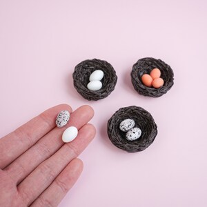 May include: Three small bird nests made of woven brown twigs. Two nests contain speckled white eggs, and one nest contains three small orange eggs.