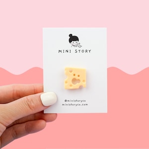 May include: A small, yellow, plastic cheese charm with holes. The charm is on a white card with the text "MINI STORY" and the social media handle "@ministoryco" and website "ministoryco.com".