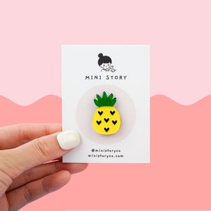 May include: A yellow pineapple enamel pin with black hearts on a white card with the text "MINI STORY" and the social media handle "@ministoryco" and website "ministoryco.com".