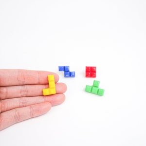 May include: Four colorful Tetris-shaped blocks, one yellow, one blue, one red, and one green. The yellow block is being held in a hand.