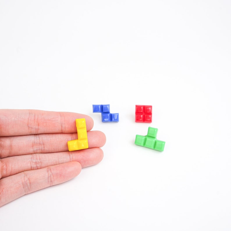 May include: Four colorful Tetris-shaped blocks, one yellow, one blue, one red, and one green. The yellow block is being held in a hand.