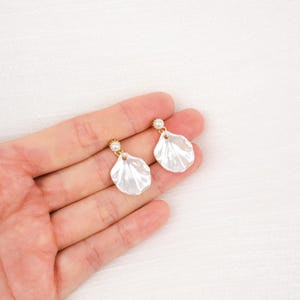 Peut inclure: Une paire de boucles d'oreilles blanches en forme de coquillage avec un accent de perle et des attaches dorées. Les boucles d'oreilles sont présentées sur un fond blanc.