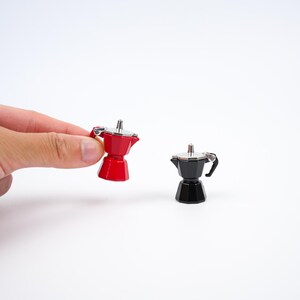 May include: Two miniature espresso makers, one red and one black, with silver accents. The red one is held by a hand. The espresso makers are small, likely for decorative purposes or as a novelty item. The background is plain white.