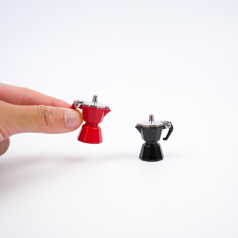 May include: Two miniature espresso makers, one red and one black, with silver accents. The red one is held by a hand. The espresso makers are small, likely for decorative purposes or as a novelty item. The background is plain white.