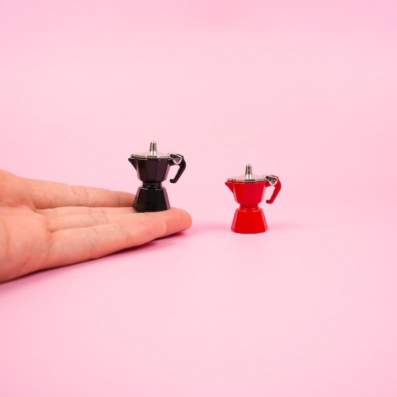 May include: Two miniature espresso makers, one black and one red, sit on a pink background. The espresso makers have silver accents and small handles. The black espresso maker is held in a hand.