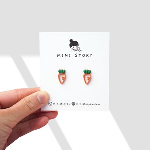 Op de afbeelding: Een paar oorbellen in de vorm van een wortel op een witte kaart met de tekst "MINI STORY" en social media-handvatten. De oorbellen zijn oranje met groene toppen.
