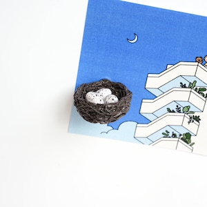 May include: A brown bird's nest with three white speckled eggs inside. The nest is sitting on a blue and white illustration of a building with a crescent moon in the sky.