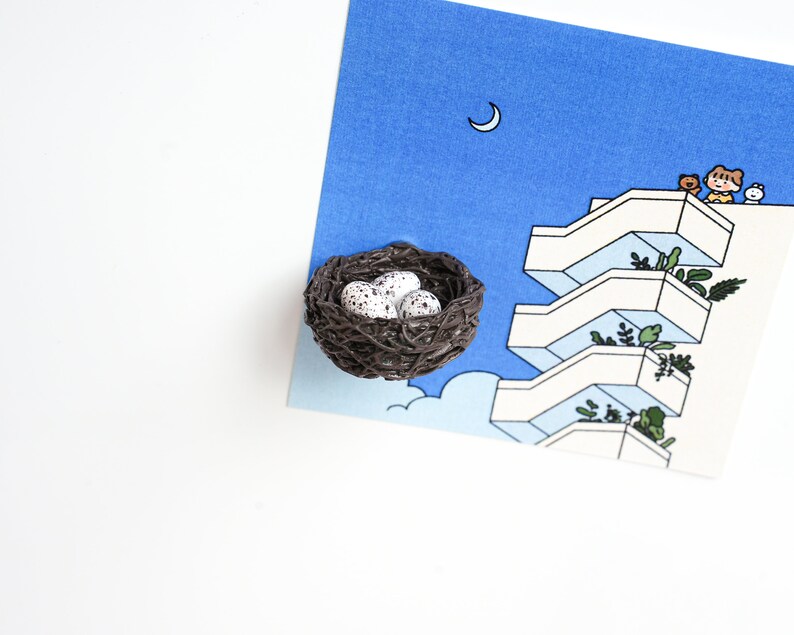 May include: A brown bird's nest with three white speckled eggs inside. The nest is sitting on a blue and white illustration of a building with a crescent moon in the sky.