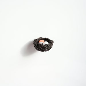May include: A small brown woven bird's nest with three speckled eggs inside. Two of the eggs are white and one is light pink.