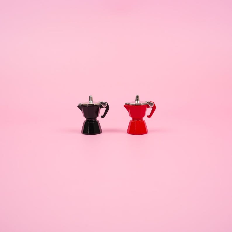 May include: Two small espresso makers, one black and one red, are displayed against a soft pink background. Each has a silver top and a black handle. The coffee makers are a classic design, perfect for brewing espresso.