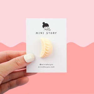 May include: A white card with the text "MINI STORY" and a cartoon illustration of a person. A small, white, half-moon shaped dumpling is attached to the card. The card is being held by a hand.
