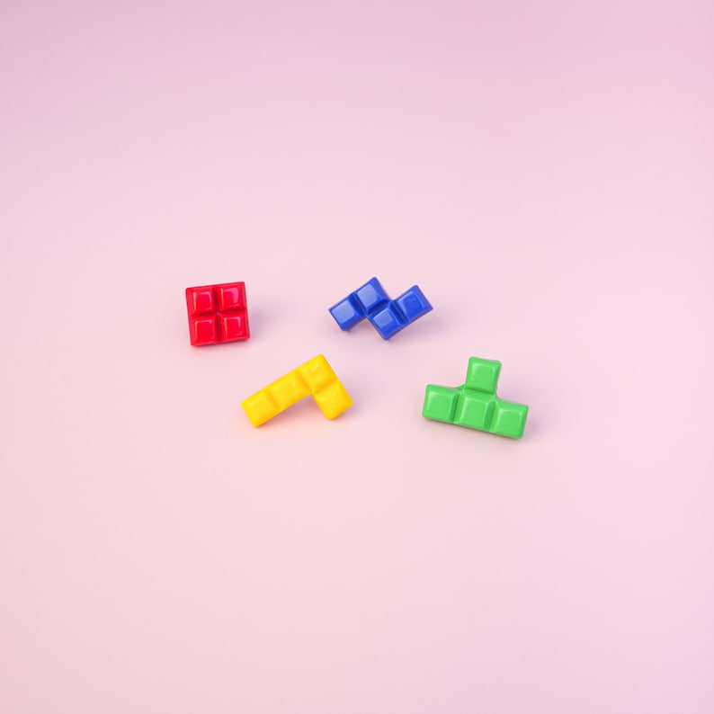 May include: Four colorful plastic Tetris blocks on a pink background. The blocks are red, blue, yellow, and green.